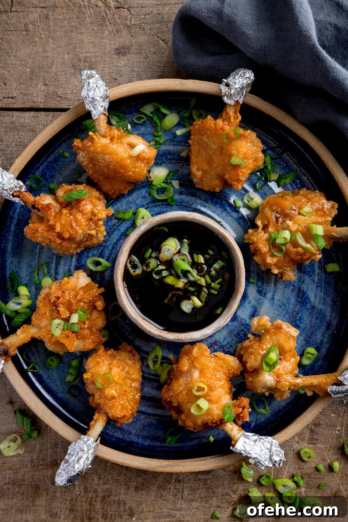 Crispy Chicken Lollipops Recipe: Finger-Licking Party Snack 5 Chicken lollipops arranged in a circle on a blue plate with a dipping bowl of sticky spicy sauce int he middle. The lollipops and sauce have been sprinkled with chopped spring onions and the plate is in a wooden table next to a blue napkin.