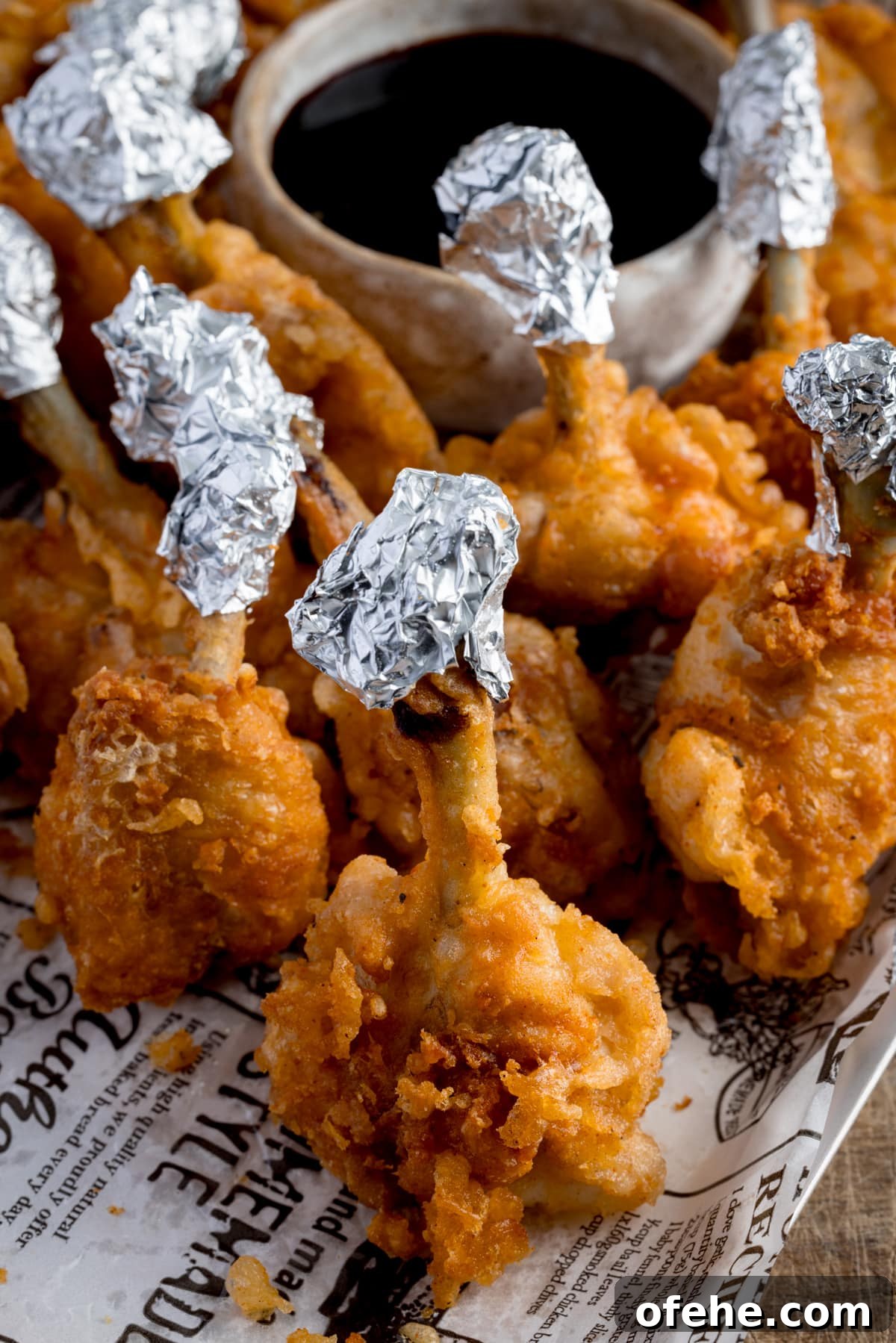 Crispy Chicken Lollipops Recipe: Finger-Licking Party Snack 6 Chicken lollipops, standing up on a piece of white baking paper that's decorated with newspaper-style writing. The lollipops have been wrapped in foil at the ends and they're next to a small light bowl of a dark dipping sauce.