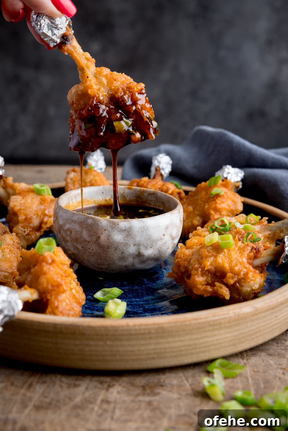 Crispy Chicken Lollipops Recipe: Finger-Licking Party Snack 7 A plate of chicken lollipops with a bowl of spicy dipping sauce. on a wooden table. One of the wings is being dipped into the sauce and is dripping. The wing is being held by fingers with pink-painted fingernails.