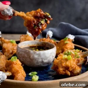Crispy Chicken Lollipops Recipe: Finger-Licking Party Snack 8 Square image of a plate of chicken lollipops with a bowl of spicy dipping sauce. One of the wings is being dipped into the sauce and is dripping. The wing is being held by fingers with pink-painted fingernails.