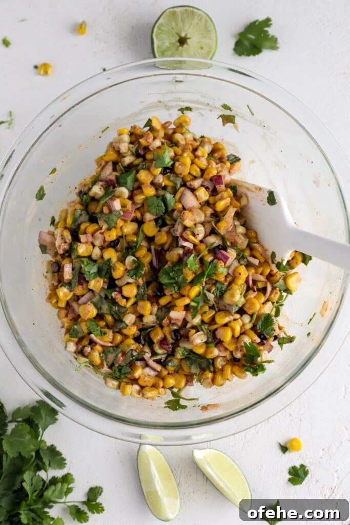 Vegan Elote-Style Corn Salad Recipe for Summer BBQs 8 Vegan Elote Corn Salad mixed with dressing in large glass bowl with spatula and lime slices on side