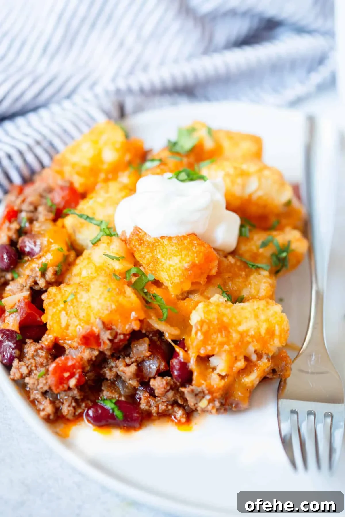 A scoop of Chili Tater Tot Casserole on a white plate with a silver fork next to it. The scoop is topped with a dollop of sour cream and a sprinkle of parsley.