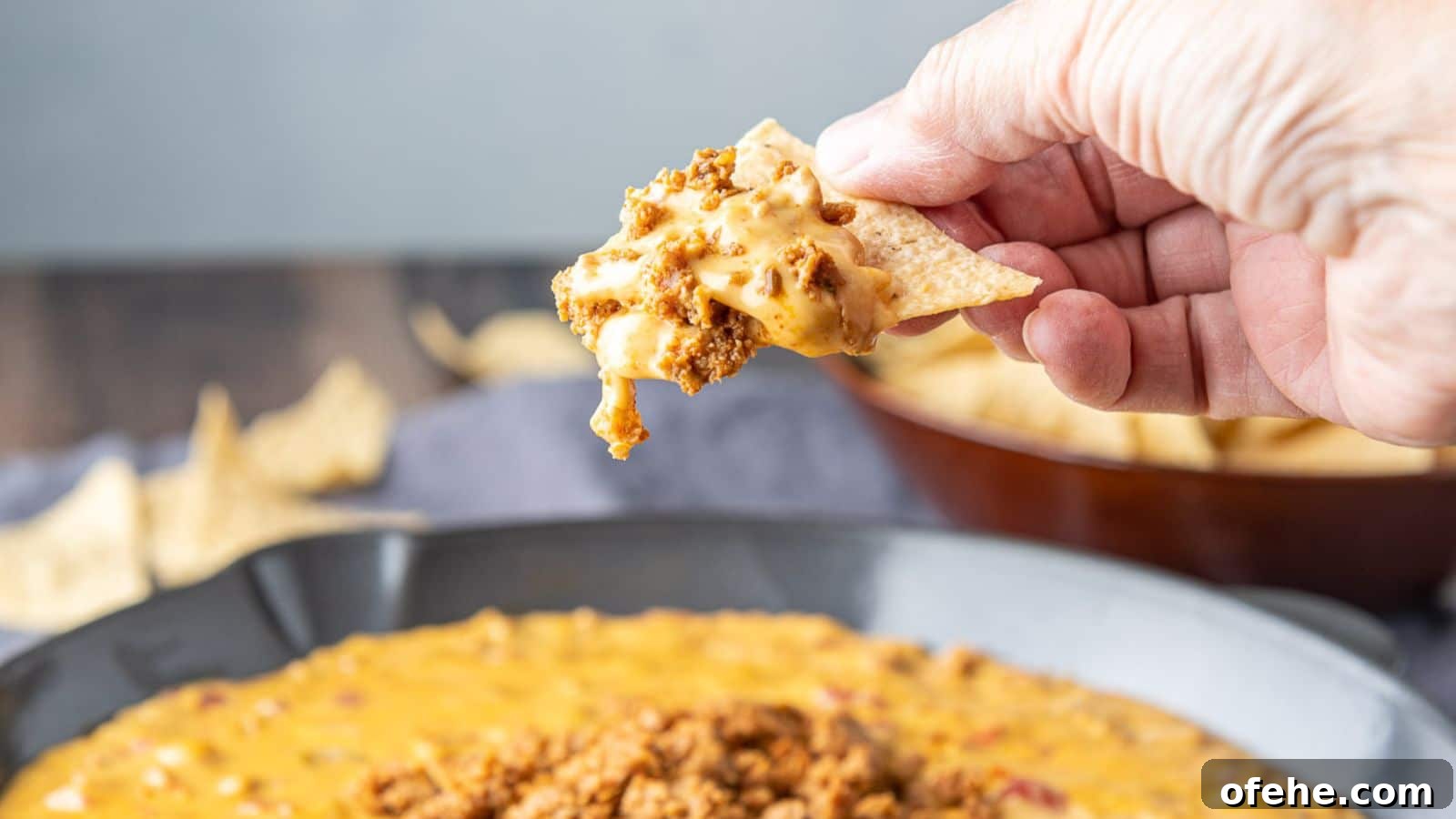 Creamy Chorizo Dip Recipe for Game Day or Parties 3 A hand holding a triangle tortilla chip filled with a cheese dip with chorizo