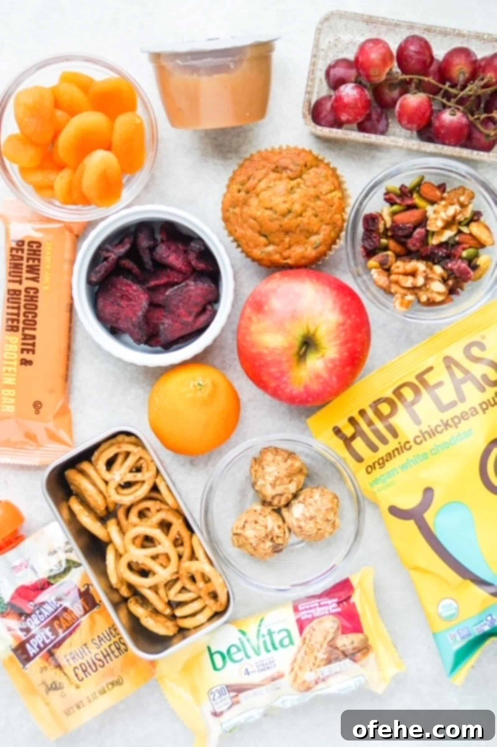 Individual snacks laid out on a surface: dried apricots, applesauce, grapes, beet chips, muffin, apple, trail mix, protein bar, spelt pretzels, HIPPEAS, energy bites, belVita package, and a apple-carrot fruit crusher from Trader Joe's.