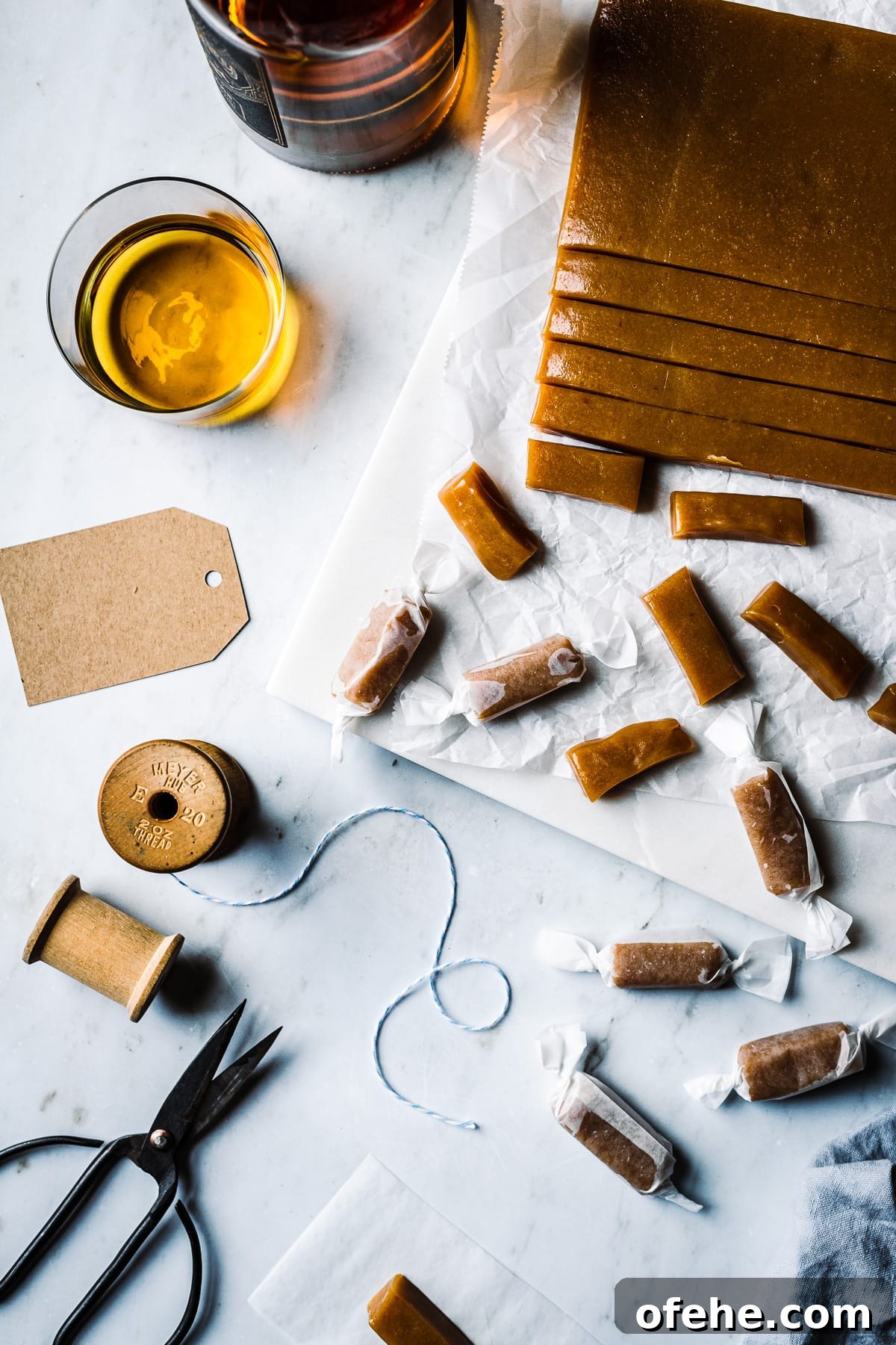 Pieces of candy being cut into individual pieces and wrapped. The work surface is grey marble. There are brown paper gift tags, a vintage spool of twine, scissors and a glass of whiskey nearby. A bottle of whiskey peeks into the frame at top left.