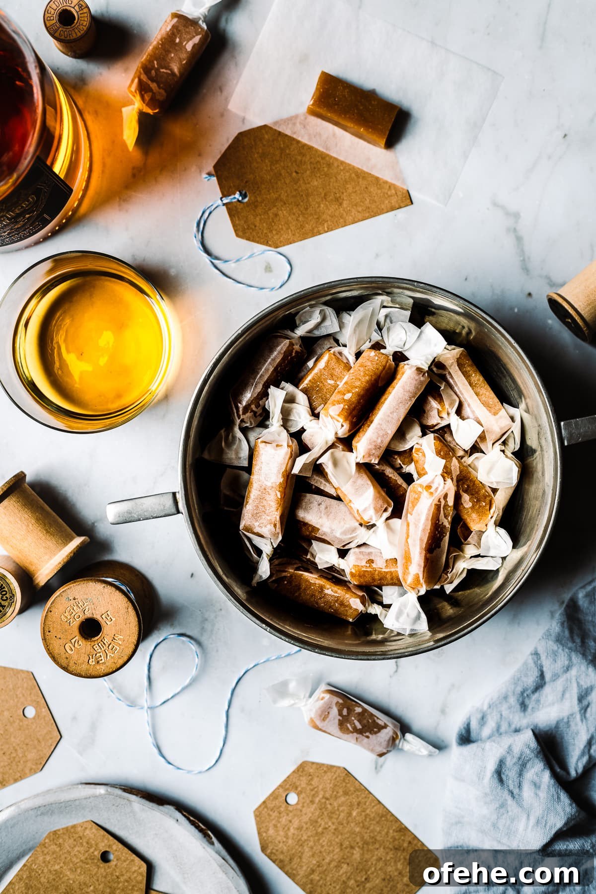 Pieces of candy wrapped in paper in a vintage silver bowl. The work surface is grey marble. There are paper candy wrappers, a vintage spool of twine, a blue linen napkin and gift tags nearby. Light shines through a whiskey bottle and glass.