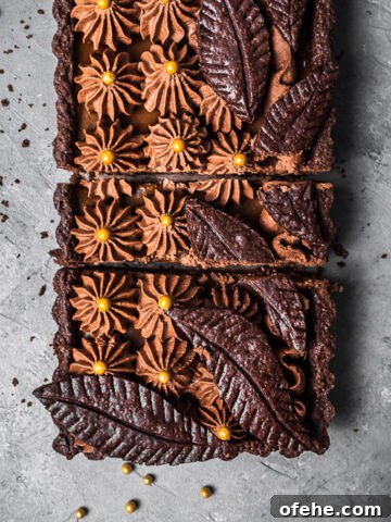 A rectangular chocolate tart with piped chocolate over layers of caramel in a chocolate pastry shell. There are decorative chocolate pastry leaves on top. It is resting on a grey textured surface.