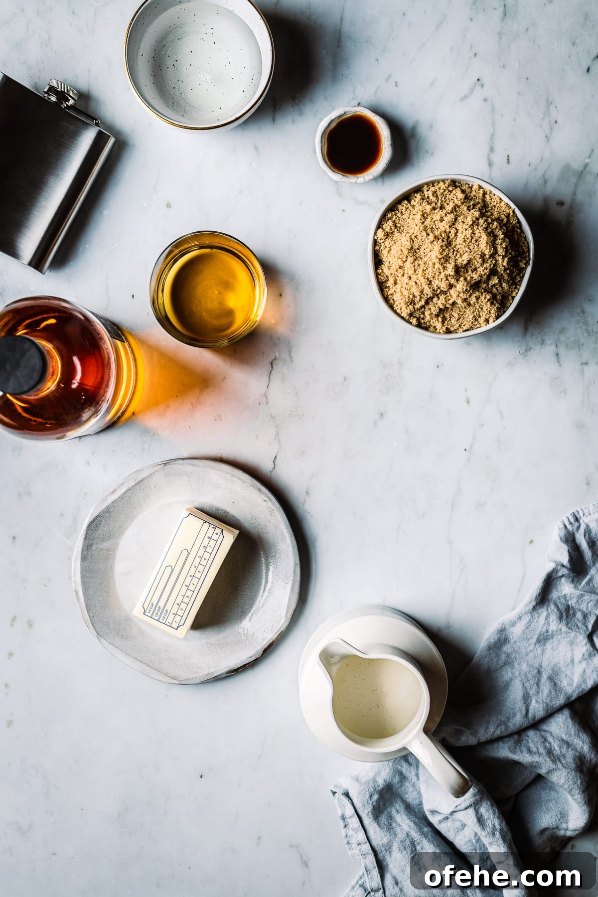 Ingredients for making caramels in containers on a grey marble surface. Light shines through a bottle of whiskey poured into a glass. Bowls of sugars and vanilla rest nearby along with a plate of butter and a pitcher of cream.