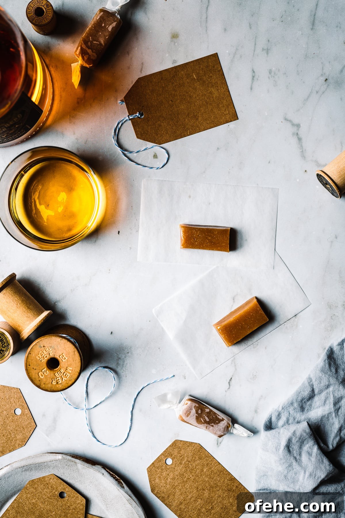 Whiskey salted caramels being wrapped on a grey marble surface. There is a vintage spool of twine, brown paper gift tags, and a blue linen napkin nearby.