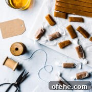 Pieces of candy being cut into individual pieces and wrapped. The work surface is grey marble. There are brown paper gift tags, a vintage spool of twine, and scissors nearby. A glass of whiskey peeks into the frame at top left.