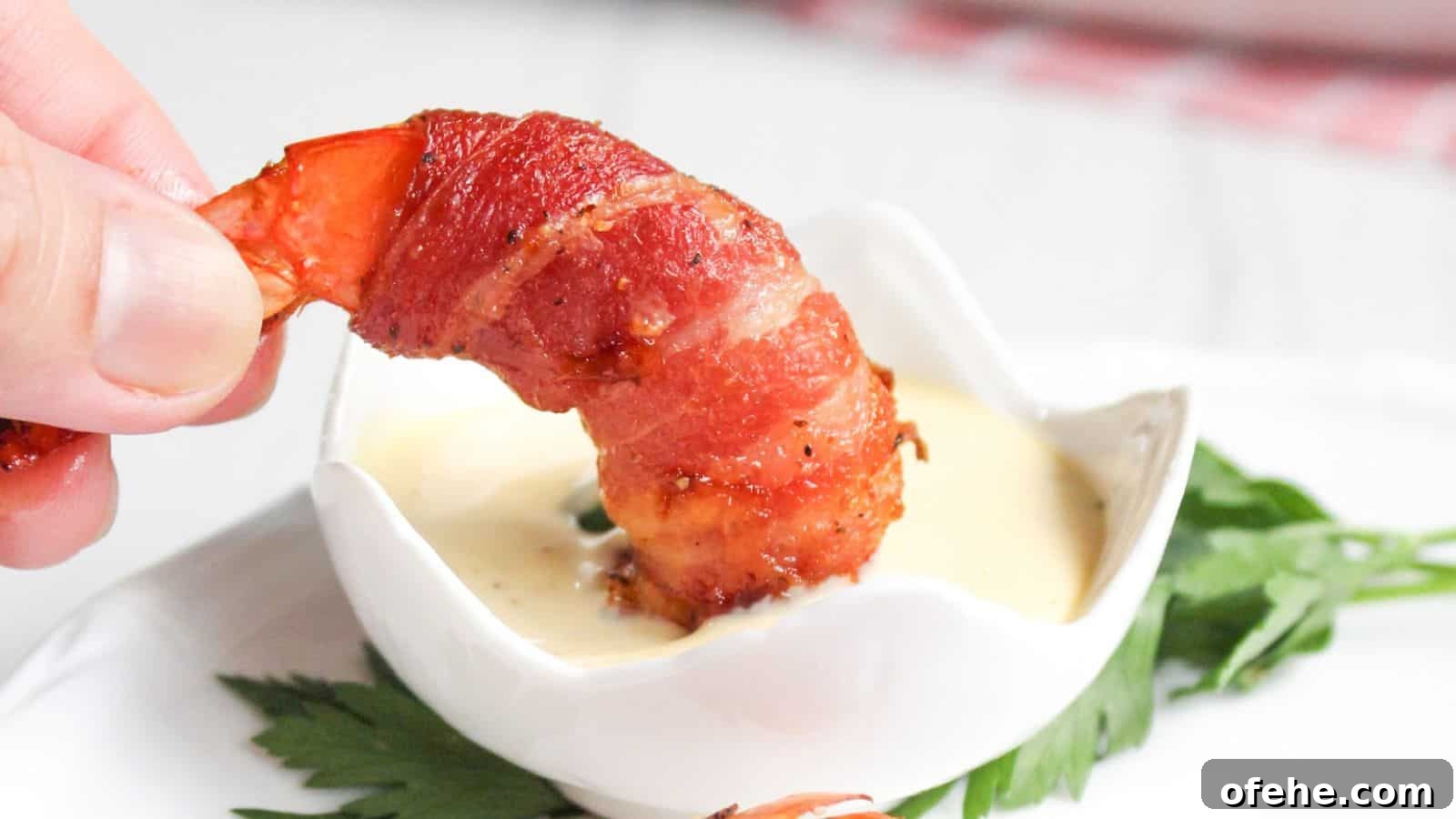 30 Quick Appetizers to Impress Guests Without Fuss 14 A hand holding a bacon-wrapped shrimp dipped in a small bowl of sauce, with a green leaf garnish on the side.