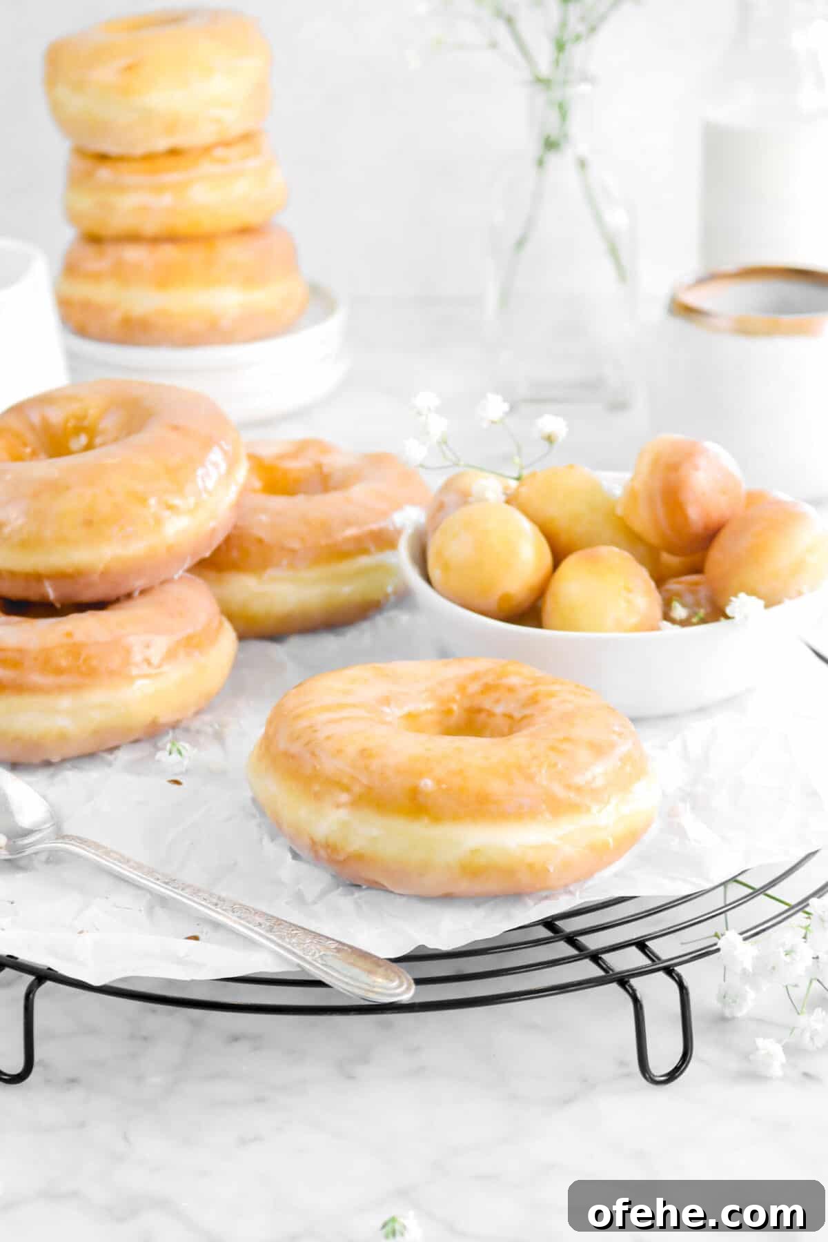 glazed doughnuts on wire cooling rack with doughnut holes in a white bowl, flowers, a stack of doughnuts behind