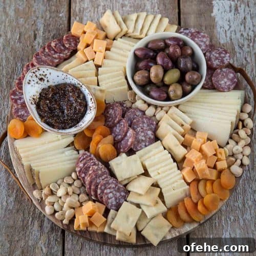 round cheese board with sliced cheese, salami, nuts, dried fruit, and little bowls of jam and olives.