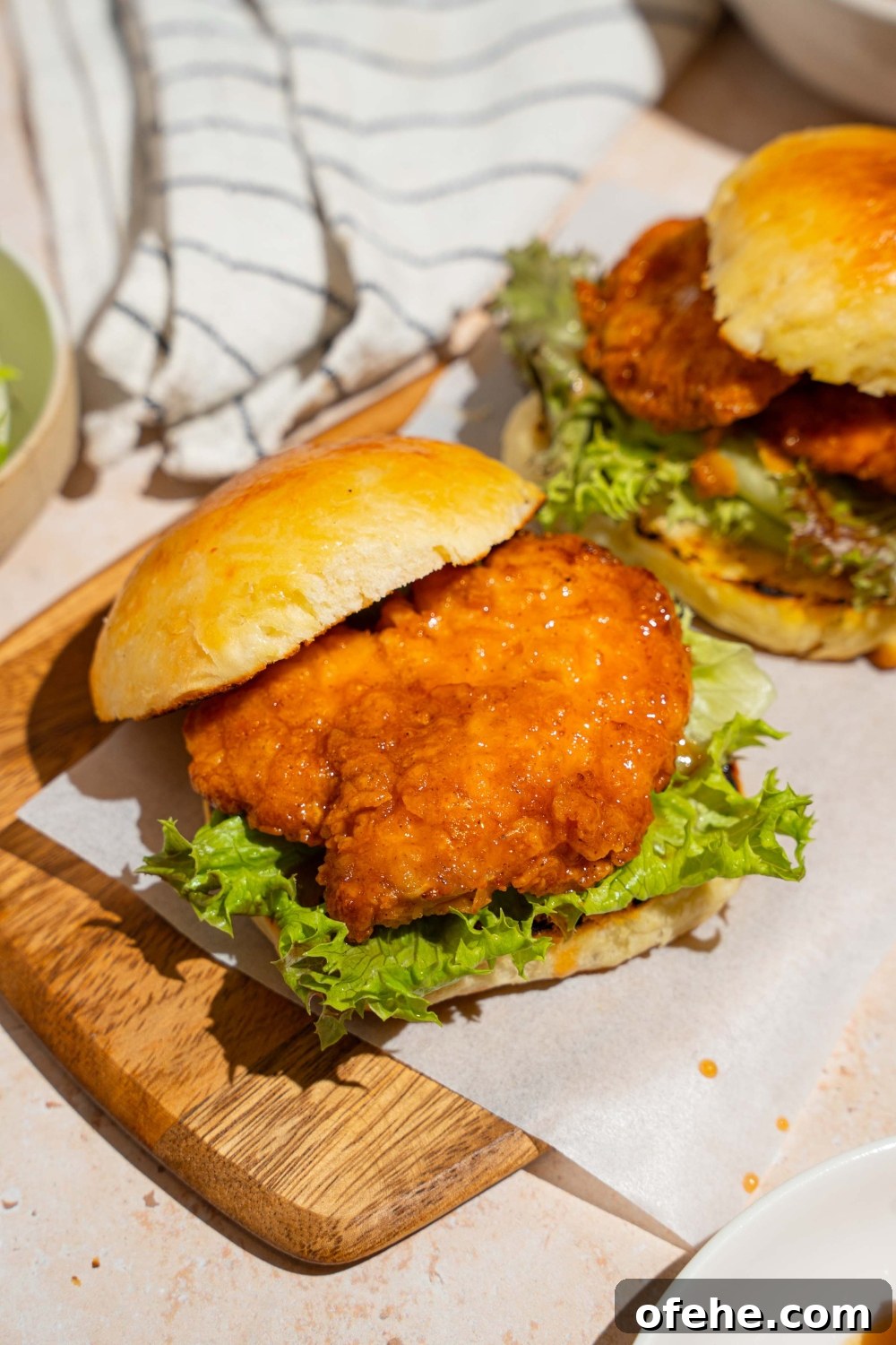 Spicy Honey Chicken Sliders with Tangy Slaw 11 A wooden board lined with parchment paper with two hot honey chicken sandwiches. The board is on a tan counter with a white striped napkin.