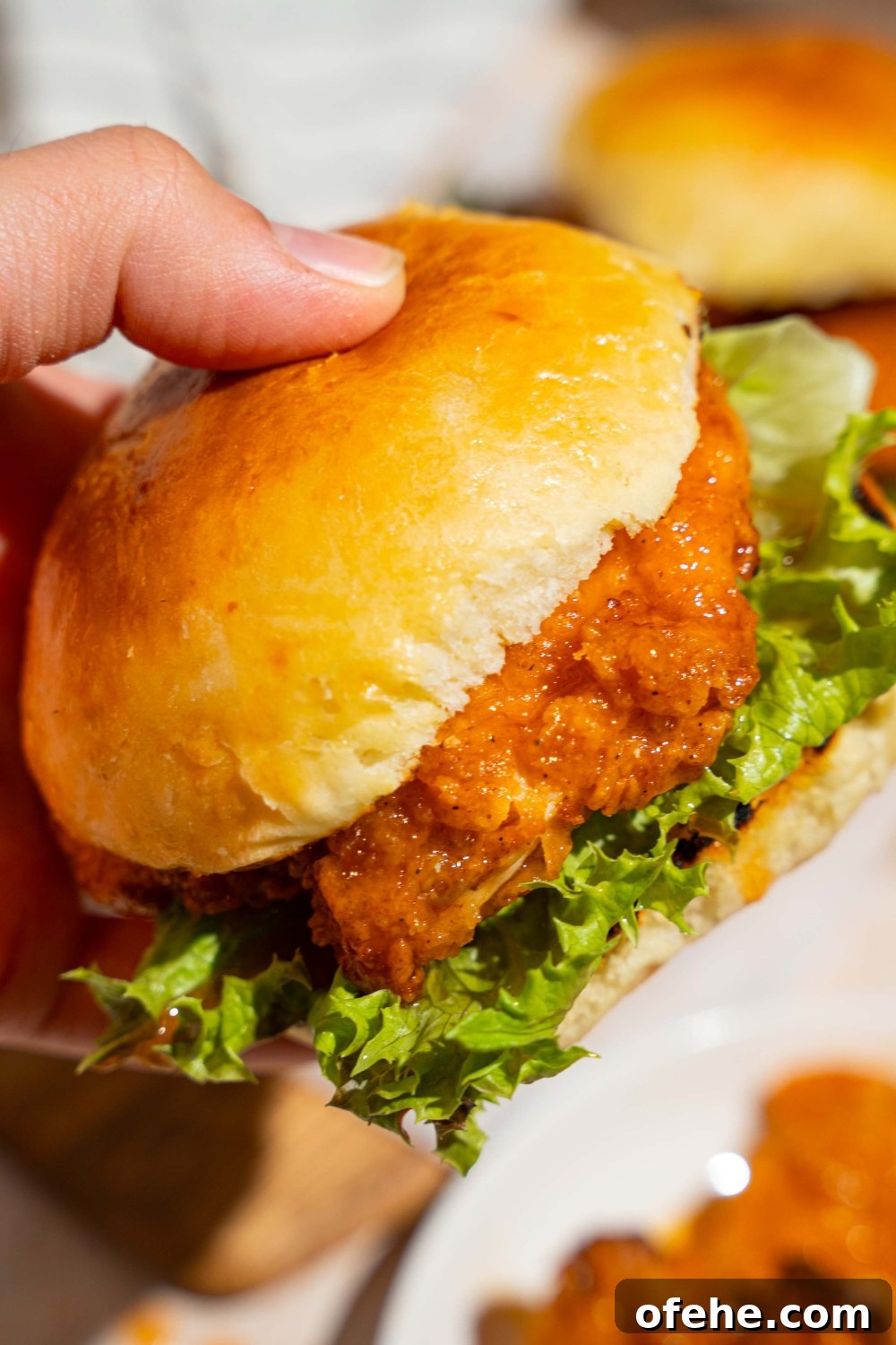 Spicy Honey Chicken Sliders with Tangy Slaw 12 A close up of a hand holding a hot honey chicken sandwich with lettuce. There is a board of sandwiches blurred in the background.