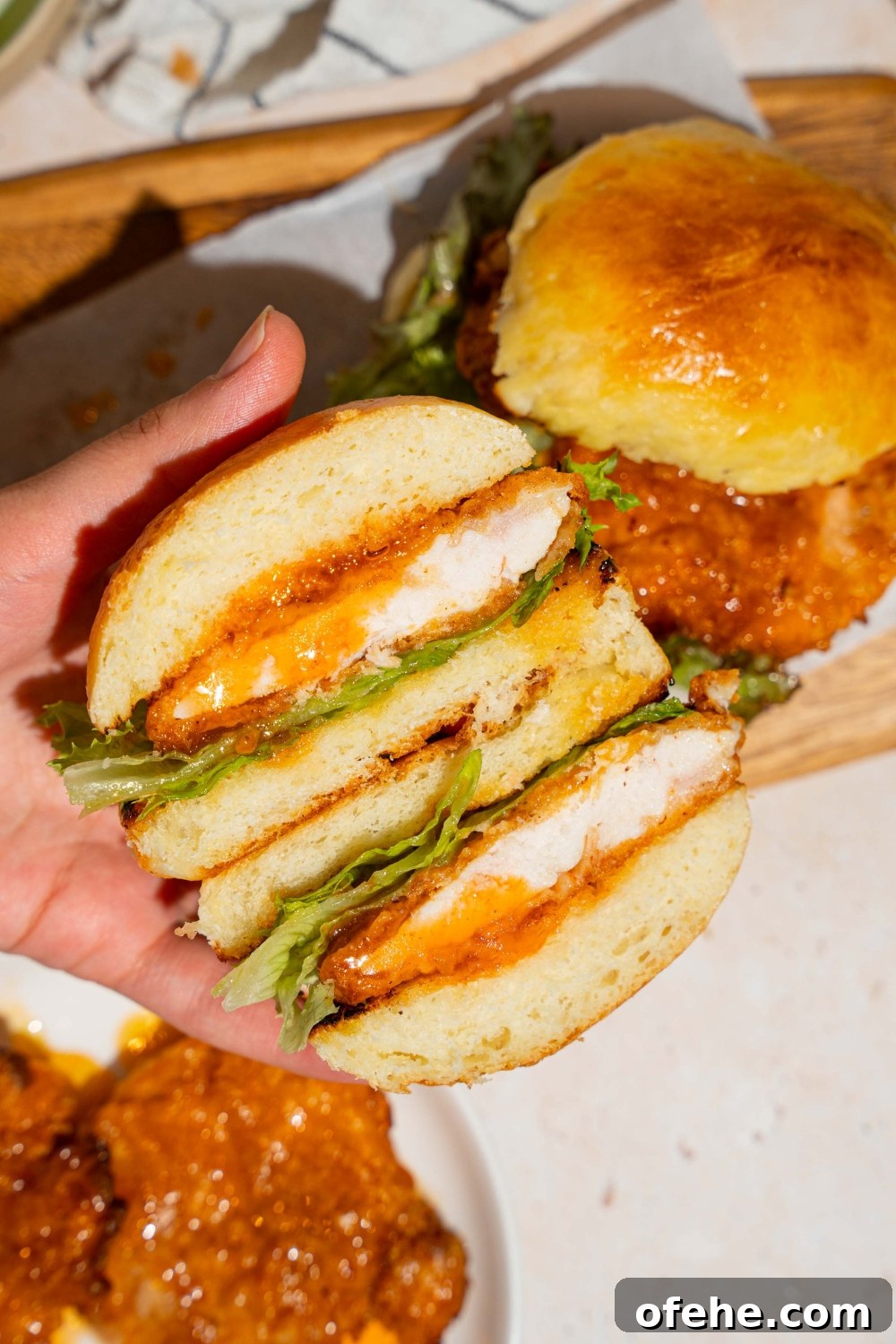 Spicy Honey Chicken Sliders with Tangy Slaw 13 A hand holding a hot honey chicken sandwich sliced in half. There is a wooden board with an additional sandwich in the background.
