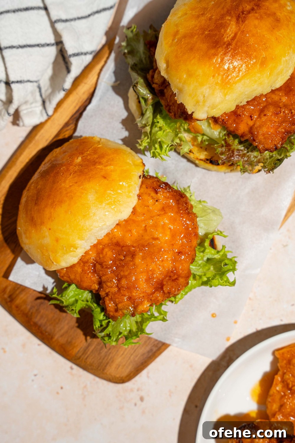 Spicy Honey Chicken Sliders with Tangy Slaw 14 A wooden board lined with parchment paper with two hot honey chicken sandwiches. The board is on a tan counter with a white striped napkin.