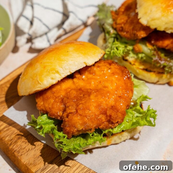 A wooden board lined with parchment paper with two hot honey chicken sandwiches. The board is on a tan counter with a white striped napkin.