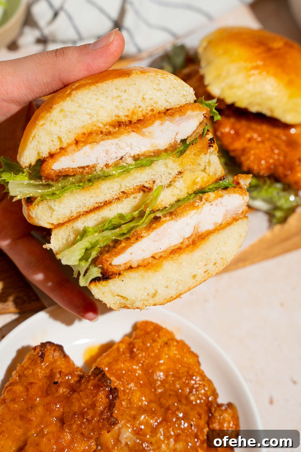 Spicy Honey Chicken Sliders with Tangy Slaw 3 A hand holding a hot honey chicken sandwich sliced in half. There is a board of sandwiches and plate of hot honey chicken in the background.