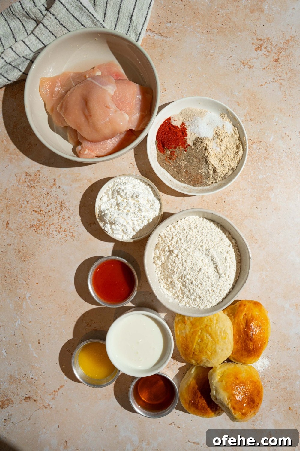 Spicy Honey Chicken Sliders with Tangy Slaw 4 An overhead shot of several bowls in various sizes containing ingredients to make hot honey chicken sandwiches including chicken breasts, buns, buttermilk, hot sauce, honey, butter, cornstarch, flour, and seasonings.