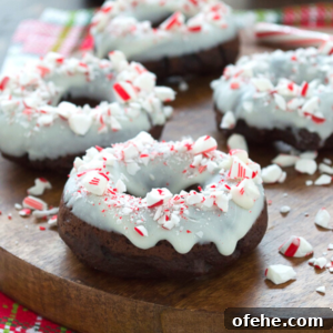 Chocolate Candy Cane Donuts: Peppermint Chocolate Donut Recipe 6 Chocolate donuts topped with white icing and crushed peppermint candies are arranged on a wooden board. Some candy cane donut pieces and peppermint bits are scattered around, creating a festive appearance.