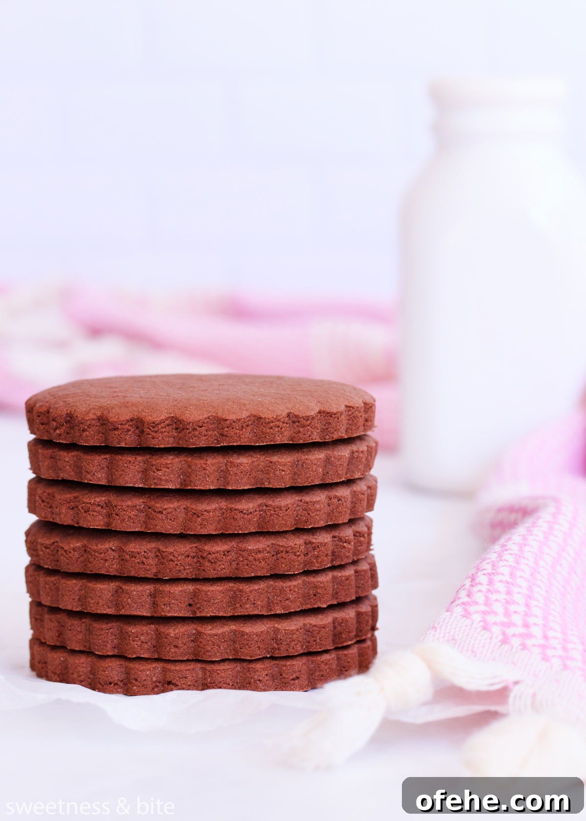 A stack of scalloped-edge round gluten free chocolate cookies, with a purple cloth and a white milk bottle.