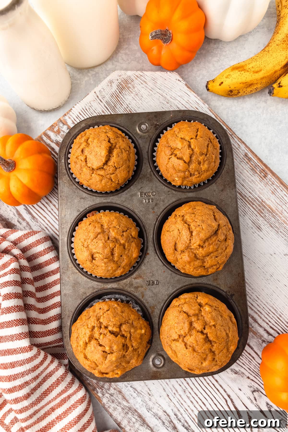 Banana Pumpkin Muffins with Warm Spices — Moist Fall Recipe 5 Cupcakes in a baking tin on a rustic wooden surface with pumpkins, bananas, and a striped cloth.