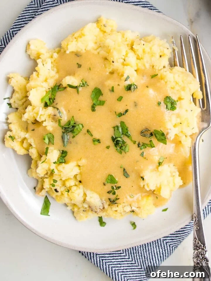 White plate filled with mashed potatoes, smothered in golden vegan gravy with fresh herbs sprinkled on top.